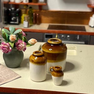 May include: A miniature kitchen scene with a green vase of pink and peach flowers, a newspaper, and three brown and white ceramic jars. The jars are different sizes and shapes, one with a handle.