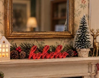 May include: A red glittery script sign reading hohoho centered on a mantelpiece. It is flanked by a white house-shaped lantern, pine branches with pinecones and red berries, a small flocked artificial Christmas tree, and a gold deer.