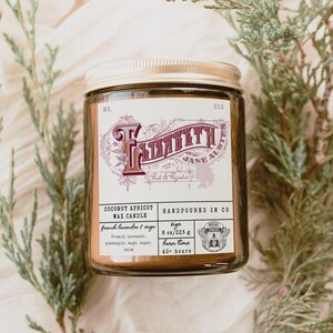 May include: A brown glass candle jar with a gold lid. The label is white with black and red text. The candle is labeled "Coconut Apricot Wax Candle" and has a burn time of 40 hours. The candle is hand-poured in the USA.