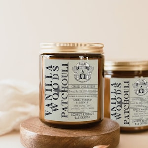 May include: Two amber glass candle jars with gold lids. The labels are white with black text and a logo. The text on the labels reads "Vanilla Woods & Patchouli", "Classic Collection", "Scent No. 24", "Coconut Apricot Wax Candle", and "Regal Embers".