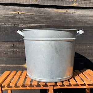 May include: A galvanised metal bucket with two handles. The bucket is cylindrical with a wide opening and sits on a wooden slatted bench. The background is a dark wooden wall.