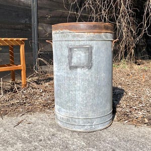 Può includere: Un bidone della spazzatura alto in metallo zincato con bordo e manici color marrone ruggine. Il bidone ha un'indentazione rettangolare sul davanti. Una panca di legno è visibile sullo sfondo.