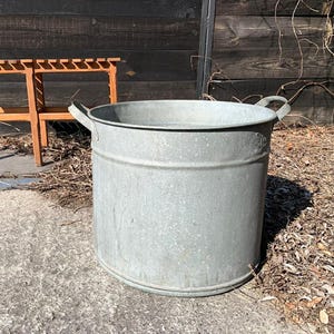 Może przedstawiać: Duża, ocynkowana metalowa wanna z dwoma uchwytami, ustawiona na betonowej powierzchni. W tle drewniana ławka z listwowym blatem. Wanna jest cylindryczna i wydaje się być w stylu vintage.