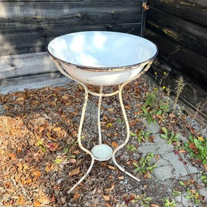 May include: A white enamel wash basin with a dark rim, supported by a cream-coloured metal stand featuring three curved legs and a small circular base plate. The basin is round and deep, resting securely within the metal frame structure.