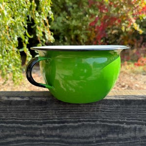May include: A bright green enamel chamber pot with a glossy finish, featuring a black handle and a black rim. The interior of the bowl is white, and the exterior is a solid, vibrant green color with a smooth, reflective surface.