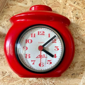 Könnte beinhalten: Eine leuchtend rote Quarzuhr mit rundem Zifferblatt und schwarzem Rahmen. Das Zifferblatt ist weiß mit schwarzen Zeigern und roten Ziffern. Auf dem Zifferblatt stehen das Wort "QUARTZ" und der Markenname "ARUL".