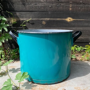 May include: A teal enamelware pot with black handles. The vintage cooking pot has a silver rim and is set against a weathered wooden background. The pot is round and appears to be in good condition.