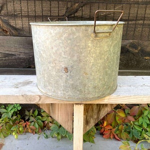 May include: A galvanised metal tub with two handles sits on a wooden bench. The tub is weathered and has a rustic look.