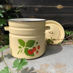 May include: A vintage cream-colored enamelware cooking pot with a matching lid. The pot features a strawberry design with green leaves and red berries. It has two side handles and a white rim. The lid is placed next to the pot. The pot is approximately 25 cm in diameter.