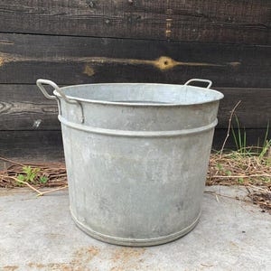 Könnte beinhalten: Eine große, verzinkte Metallwanne mit zwei Griffen. Die Wanne ist zylindrisch mit einer weiten Öffnung und einem leicht konischen Boden. Das Metall hat ein verwittertes, graues Aussehen. Der Hintergrund ist eine dunkle Holzwand.