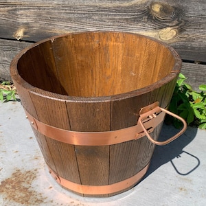 Könnte beinhalten: Ein Holztopf mit einem Kupferband und einem Griff. Der Topf ist braun und hat eine natürliche Holzoptik.