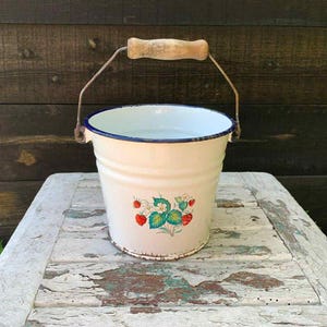 Könnte beinhalten: Cremefarbener Emaille-Eimer mit blauem Rand und Holzgriff. Der Eimer hat ein Erdbeermuster. Der Griff ist aus Metall und Holz. Der Eimer steht auf einem weißen, abgenutzten Tisch.