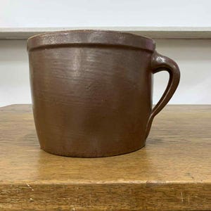 Może przedstawiać: Brązowy ceramiczny kubek z uchwytem. Kubek ma cylindryczny kształt i lekko rozszerzany brzeg. Zewnętrzna strona ma teksturowany wygląd. Kubek stoi na drewnianej powierzchni.