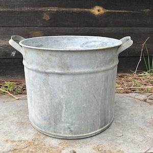 Op de afbeelding: Een verweerde, gegalvaniseerde metalen emmer met twee gebogen handvatten. De emmer is cilindrisch met een licht uitlopende rand en staat op een betonnen oppervlak. De achtergrond heeft een donkere houten muur en wat gebladerte.
