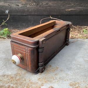 Könnte beinhalten: Eine Vintage-Holzschublade mit dunkelbraunem Finish. Die Schublade hat einen weißen und roten Knopf mit Messingakzenten. Die Schublade hat dekorative vertikale und horizontale Linien auf der Vorderseite und dekorative Säulen.