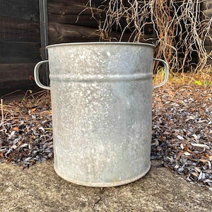 May include: A large, weathered metal bucket with two curved handles. The cylindrical container has a slightly textured surface and a silver-grey colour. It is outdoors, resting on a concrete surface, with a wooden structure and foliage in the background.