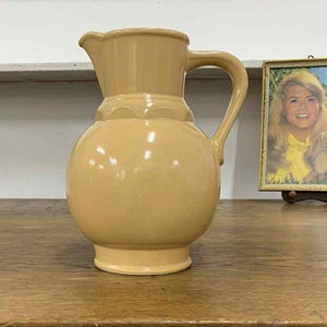 Puede incluir: Una jarra de cerámica de color beige con un cuerpo redondeado, un asa y un pico vertedor. La jarra está sobre una superficie de madera. Una fotografía enmarcada de una mujer y un perro está en el fondo.
