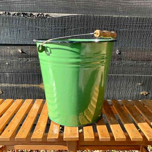 Puede incluir: Un cubo de esmalte verde con un asa de madera y un alambre de metal. El cubo es cilíndrico y está sobre un banco de madera. El fondo es una pared de madera oscura.