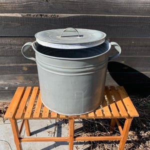 Puede incluir: Un cubo de metal galvanizado gris con tapa y dos asas. El cubo está sobre un pequeño banco de madera. La tapa está ligeramente entreabierta, revelando el interior. El fondo es una pared de madera desgastada.