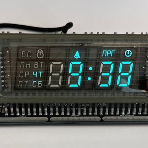 Könnte beinhalten: Eine transparente digitale Uhr aus Kunststoff mit blauen LED-Ziffern, die die Zeit 12:31 anzeigen. Die Uhr hat ein schwarzes Kabel, das an der Rückseite befestigt ist, und eine schwarze Platine mit Stiften, die mit "Datum", "Alarm", "WLAN-Signal" und "Entworfen von" beschriftet sind.