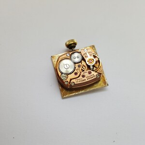 Puede incluir: La parte posterior de un reloj de pulsera dorado con una caja cuadrada. El movimiento del reloj es visible a trav&eacute;s del fondo de la caja, y el texto "OMEGA WATCH CO. SWISS MADE" est&aacute; impreso en el movimiento.