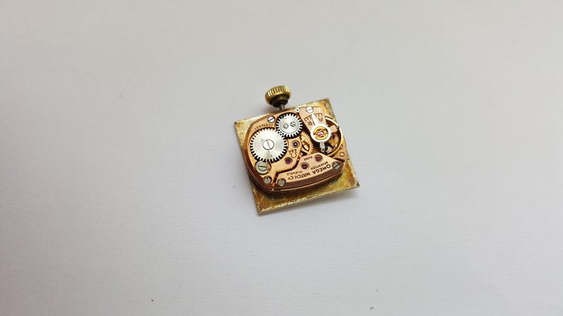 Puede incluir: La parte posterior de un reloj de pulsera dorado con una caja cuadrada. El movimiento del reloj es visible a trav&eacute;s del fondo de la caja, y el texto "OMEGA WATCH CO. SWISS MADE" est&aacute; impreso en el movimiento.