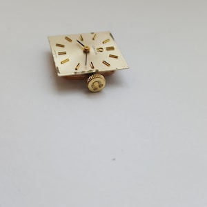 Puede incluir: Una esfera de reloj de color dorado con agujas negras y n&uacute;meros romanos. La esfera del reloj es cuadrada y tiene un peque&ntilde;o logotipo omega dorado en la parte inferior.