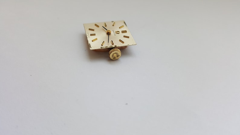 Puede incluir: Una esfera de reloj de color dorado con agujas negras y n&uacute;meros romanos. La esfera del reloj es cuadrada y tiene un peque&ntilde;o logotipo omega dorado en la parte inferior.