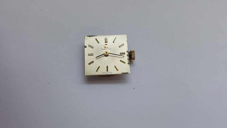Puede incluir: Una esfera de reloj de forma cuadrada con agujas y marcas doradas. La esfera del reloj es blanca con la palabra "OMEGA" impresa en negro.