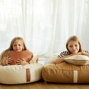 Op de afbeelding: Twee meisjes ontspannen op beige en bruine vloer kussens met kussens. De kussens hebben een bruine leren rand. De meisjes glimlachen en kijken naar de camera.