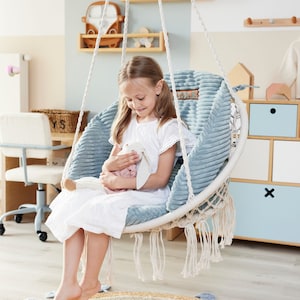 Op de afbeelding: Een jong meisje zit in een blauwe en witte hangstoel met een wit knuffelkonijn op schoot. De stoel hangt aan het plafond met een wit touw. Het meisje draagt een witte jurk en heeft haar haar in twee vlechten. De stoel hangt boven een lichtbruin tapijt met een blauw-wit gestreepte rand.