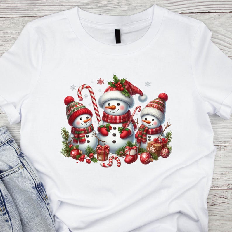 May include: White t-shirt with a Christmas design featuring three snowmen. The snowmen are wearing red and white scarves and hats. The design also includes candy canes, holly, and presents.