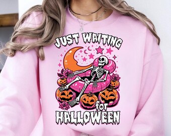 May include: Pink sweatshirt with a Halloween-themed graphic. The design features a skeleton relaxing on a pink inflatable ring, surrounded by pumpkins, a crescent moon, and stars. Text reads "JUST WAITING for HALLOWEEN."
