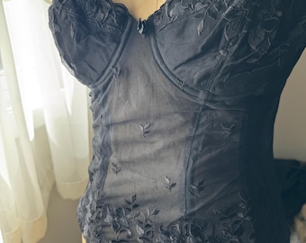May include: Black strapless corset with floral embroidery and sheer mesh panels. The corset is displayed on a vintage dress form. The top edge has a scalloped trim. The corset is a lingerie item.