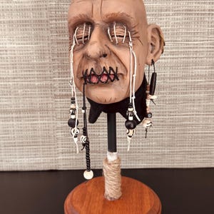 May include: A sculpted head with stitched eyes and mouth, adorned with dangling beaded accents. The head is mounted on a wooden base with a black metal rod wrapped in twine. The face has a weathered, aged appearance.