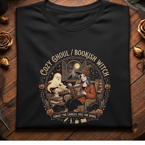 Op de afbeelding: Een zwart t-shirt met een afbeelding van een heks met een hoed die een boek leest, een geest met een bril, een kat en pompoenen. De tekst luidt COZY GHOUL / BOOKISH WITCH en FORGET THE CANDLES, PASS THE BOOKS, met een open raam en een volle maan.