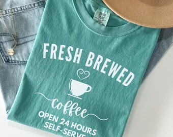 May include: A teal blue t-shirt with the text "Fresh Brewed Coffee Open 24 Hours Self-Serve" in white. A stylized coffee cup with a heart-shaped steam is above the word "coffee".