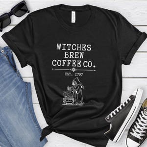 Könnte beinhalten: Schwarzes T-Shirt mit dem weißen Text "Witches Brew Coffee Co." und einer Illustration einer Hexe. Das Shirt enthält auch den Text "EST. 1797".