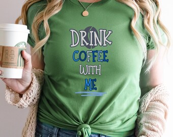 May include: A green t-shirt with the text "DRINK COFFEE WITH ME" in blue and white lettering. The shirt has a coffee bean graphic and a blue line underneath the text.