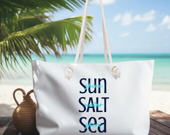 May include: A white tote bag with rope handles and the words "Sun, Salt, Sea" in navy blue and light blue. The bag is on a wooden surface with a woven basket and palm leaves in the background, with a beach scene.