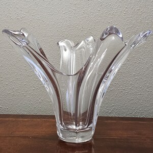 May include: Clear glass vase with a unique, flared design. The vase has a wide opening and a series of curved, petal-like edges. The glass is transparent, allowing light to pass through, and sits on a wooden surface.