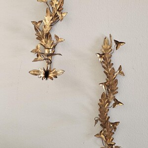 May include: Two gold-colored wall sconces with a leaf and floral design. The sconces are made of metal and have a vintage look. They are mounted on a light-colored wall. The sconces are decorative and add a touch of elegance to any room.