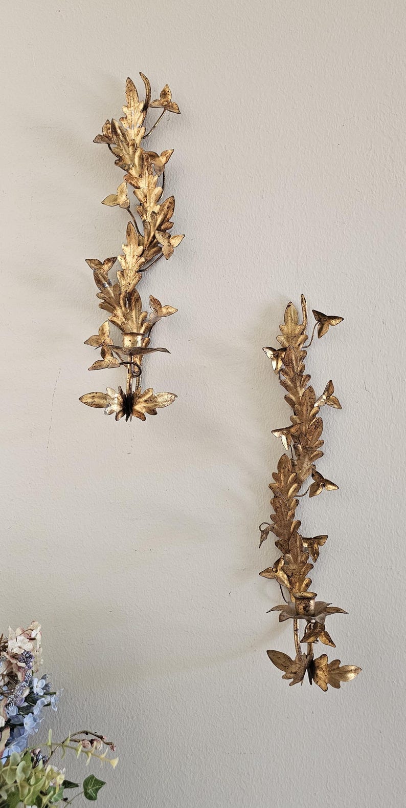 May include: Two gold-colored wall sconces with a leaf and floral design. The sconces are made of metal and have a vintage look. They are mounted on a light-colored wall. The sconces are decorative and add a touch of elegance to any room.