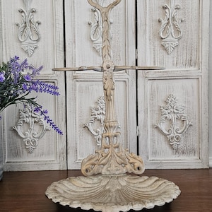 May include: An antique-style, off-white metal tiered serving stand with ornate, floral detailing. The stand features a scalloped base, a central column with crossbars, and a handle at the top. The background includes white, distressed wooden panels.