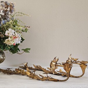 May include: Gold-colored metal decorative leaf and bird accents. The decorative pieces are arranged on a white surface. A vase of flowers is in the background. The metal pieces have a vintage aesthetic.