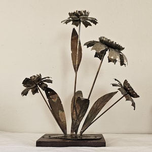 May include: Metal flower sculpture with four blooms and several leaves. The flowers are made of a dark, textured metal and are attached to thin stems. The sculpture is mounted on a dark wooden base.