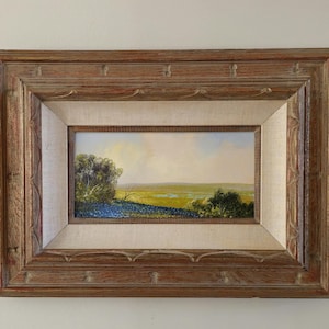 May include: An oil painting of a landscape scene, framed in a rustic wooden frame. The artwork depicts a field of blue flowers, green grass, and trees under a yellow and blue sky. The frame is a light brown color.