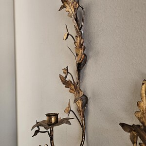 May include: A gold-colored wall sconce with a floral and leaf design. The sconce has a small cup for holding a candle. The design includes leaves and flowers, creating an elegant, decorative piece.
