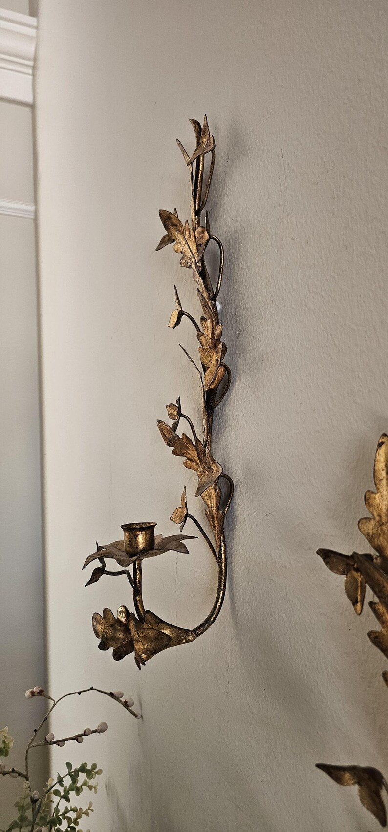 May include: A gold-colored wall sconce with a floral and leaf design. The sconce has a small cup for holding a candle. The design includes leaves and flowers, creating an elegant, decorative piece.