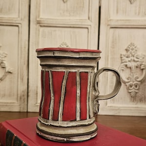 May include: A handmade ceramic mug with a red and cream color scheme. The mug features vertical red stripes and a decorative handle. The mug is sitting on a red book.